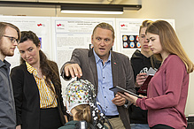 Ein Mann spricht zu vier Studierenden und zeigt auf eine Haube mit vielen Kabeln.