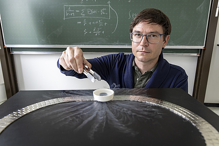 A man sits in front of a blackboard and looks into the camera. He holds a pair of pliers over a small white container.