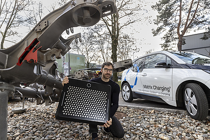 A man in front of a car and a construction part of a railway is holding a board with metal circles on it