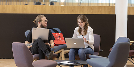 Ein junger Mann und eine junge Frau, beide mit Laptop und im Gespräch miteinander