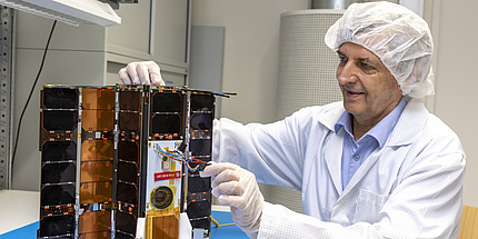 Ein Mann mit weißem Labormantel und weißem Häubchen über den Haaren sitzt an einem Schreibitsch und arbeitet an einem Satelliten. Der Satellit steht aufgeklappt mit den schwarzen Solarpanelen nach vorne auf dem Tisch.