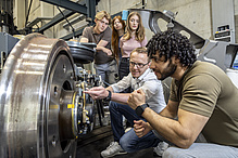 Two men are kneeling next to a railway wheel, three people are standing in the background.