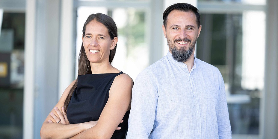 Eine Frau (links im Bild) und ein Mann (rechts im Bild) lächeln in die Kamera. Die Frau hat die Arme verschränkt, der Mann die Hände in der Hosentasche. 