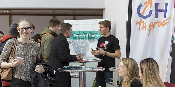 Ein junger Mann in TU Graz-T-Shirt beräte Leute an einem Stehtisch.