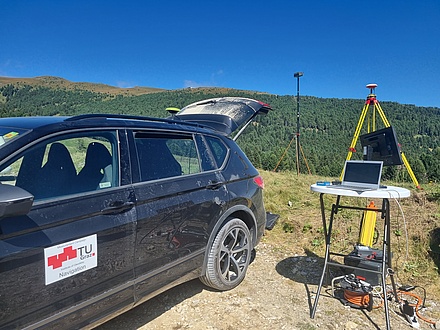 A car is parked next to a small table with a computer on it. Behind the table is a GNSS antenna.