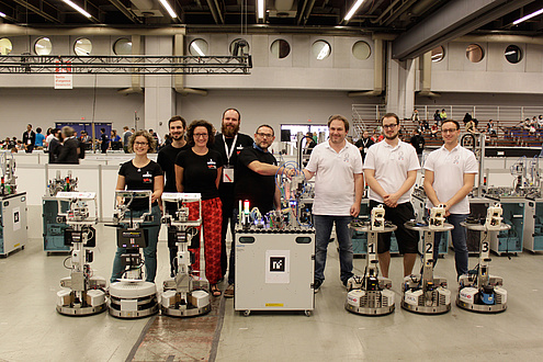 The world champion team GRIPS consisting of two women and three men in black T-shirts and the vice world champion team PYRO consisting of three men in white T-shirts posing with their sehcs robots in a production hall.