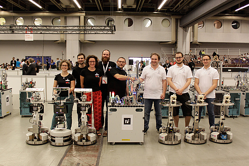 Das Weltmeisterteam GRIPS bestehend aus zwei Frauen ud drei Männern in schwarzen T-Shirts und das Vizeweltmeisterteam PYRO bestehnd aus drei Männern in weißen T-Shirts posieren mit ihren insgesamt sehcs Robotern in einer Produktionshalle