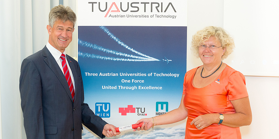 Rektor Harald Kainz und Rektorin Sabine Seidler mit einem rot-weiß-roten Staffelholz vor einem TU Austria Roll-up