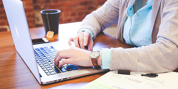 Eine Person tippt auf einer Laptop-Tastatur. Im Vordergrund ist ein Kalender, im Hintergrund sind ein Handy und eine Tasse Tee auf dem Schreibtisch zu sehen.