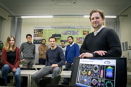 Gruppe von Menschen in Büro, rechts Bildschirm mit Abbildungen von Nanostrukturen, hinten ein Poster zu Nanoanalytik