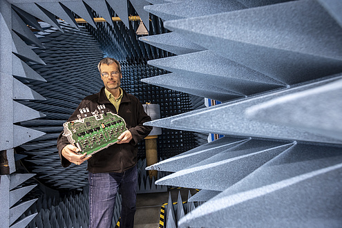 A man stands in a chamber with angular spikes protruding from the wall and holds a large electronic component in his hands.