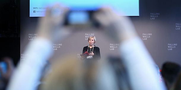 Eine Frau hält Moderationskarten in der Hand und spricht vor einer Wand, auf der mehrfach der Schriftzug "Falling Walls Lab" zu sehen ist. Im Vordergrund ist unscharf Publikum zu erkennen.