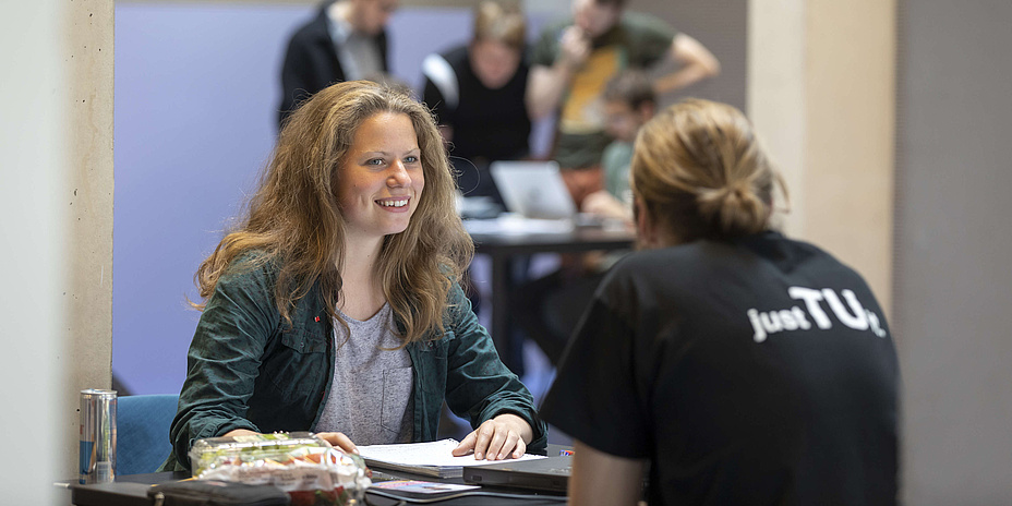 Eine junge Frau und ein junger Mann sitzen sich an einem kleinen Tisch gegenüber, auf dem ein Sandwich und Arbeitsmaterialien liegen, im Hintergrund ein Stehtisch mit drei Studierenden und einem Laptop.