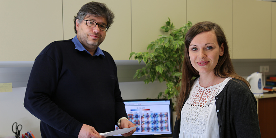 A man in a black sweater and a woman in a white blouse are smiling at the camera. There is a computer screen with colourful objects at the back.