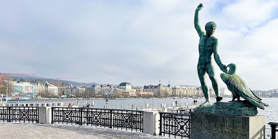 Blick über einen See. Im Hintergrund sieht man eine Stadt. Im Vordergrund eine Statue.