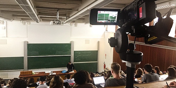 Im Vordergrund ist eine Videokamera zu sehen, die das Geschehen im Hintergrund aufzeichnet: ein Professor trägt vor einem vollen Hörsaal vor.