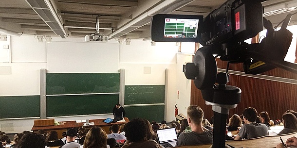 In the foreground is a video camera that records the events in the background: a professor in front of a full auditorium.