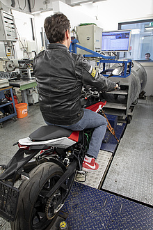 A man in a black leather jacket can be seen from behind; he is sitting on a motorcycle standing on a chassis dynamometer;