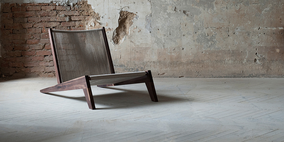 Lounge chair made of nut wood and natural coloured hemp rope against a partially coarsely plastered brick wall.