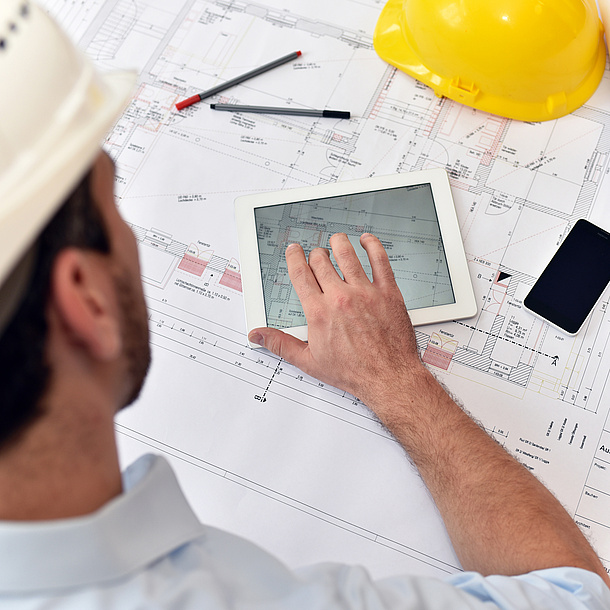 A man with a construction helmet, in front of him a table with construction plans, pens, tablet and mobile phone.