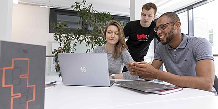 Zwei junge Männer und eine junge Frau mit Laptop und Arbeitsunterlagen an einem Tisch..