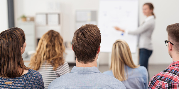 Eine Frau steht an einem Flipchart und erklärt. Im Vordergrund sind die Hinterköpfe mehrerer Studierender zu sehen, die zuhören.