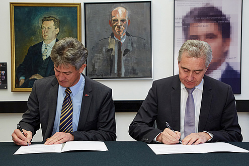 Zwei Herren im Anzug unterzeichnen an einem Tisch sitzend zwei Verträge.