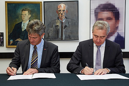 Zwei Herren im Anzug unterzeichnen an einem Tisch sitzend zwei Verträge.