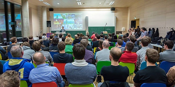 Viele Menschen sitzen in einem Hörsaal und hören einem Vortragendem zu.