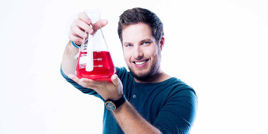 Junger Mann mit Bart und blauem Langarmshirt hält ein Messgefäß mit knallrotem Inhalt mit beiden Händen auf Augenhöhe.