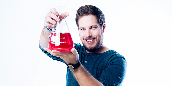 Junger Mann mit Bart und blauem Langarmshirt hält ein Messgefäß mit knallrotem Inhalt mit beiden Händen auf Augenhöhe.