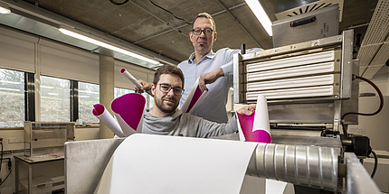 Zwei Männer stehen hinter einer großen Rolle Papier und schauen in ide Kamera.