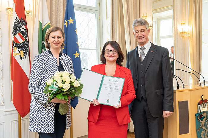 Eine Frau mit Blumen, eine mit Urkunde und ein Mann posieren vor Österreich- und EU-Fahnen