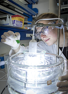 Close-up of a woman wearing laboratory glasses looking at a small bottle of green liquid over a light source.