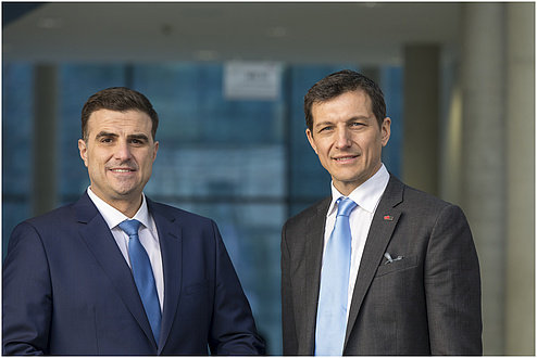 Two smiling gentlemen. The gentleman on the left is wearing a blue suit with a light blue tie. The gentleman on the right is wearing a grey suit with a light blue tie.