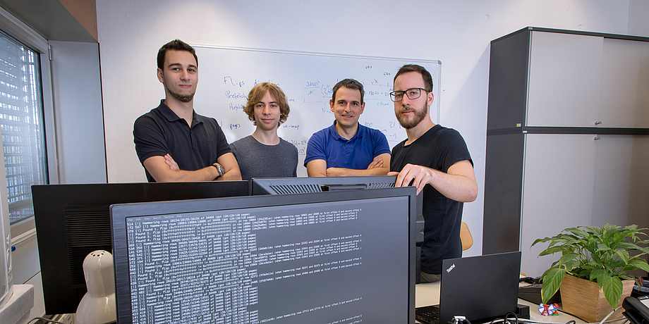 Four man are standing behind a black computer screen with white letters on it.