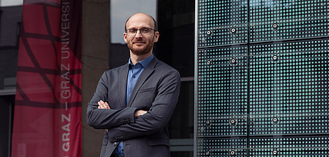 Ein Mann steht mit verschränkten Armen vor einem Gebäude und schaut in die Kamera.