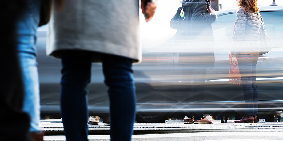 Wartende und gehende Personen vor und auf einem Zebrastreifen mit vorbeiziehenden Autos.