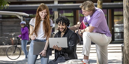Three students look at an open laptop computer while chatting at the campus.