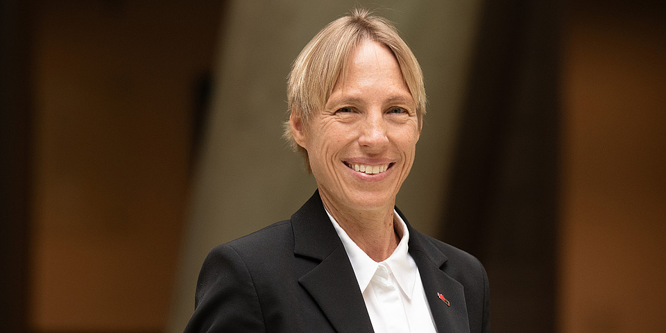 A woman is standing in a hallway and is looking into the camera with a smile.