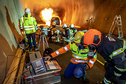 Several people in protective clothing with measuring devices, fire behind them.