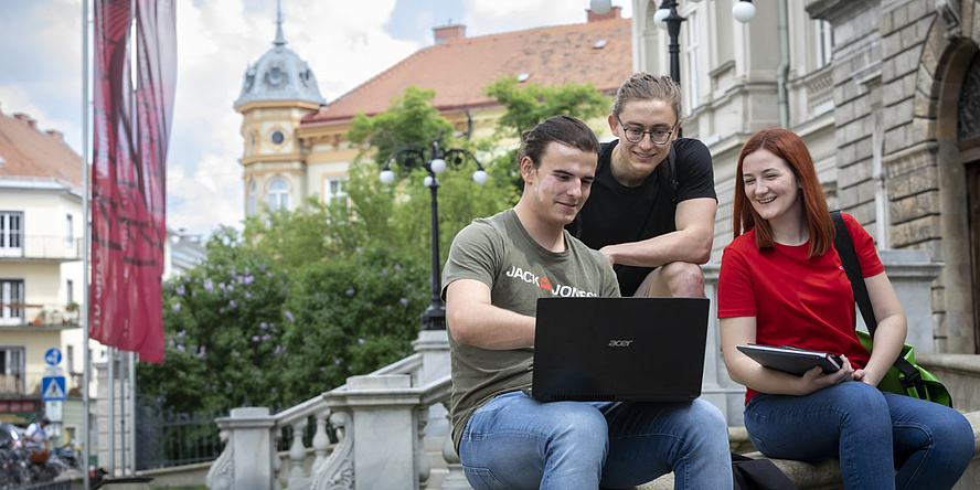 Drei Personen sitzen auf einem Treppenasatz und schauen auf den Bildschirm eines Laptops.
