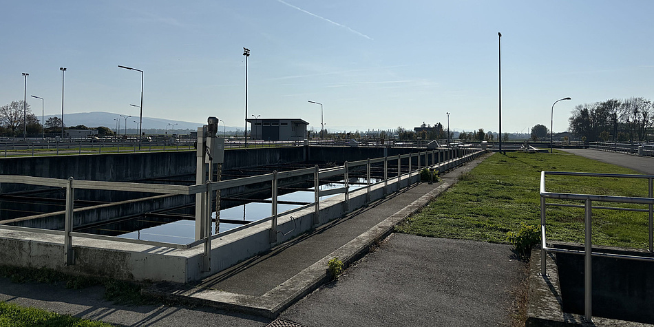 Photo of a sewage treatment plant. The sky is blue, and below it, on the left, there is a basin with a railing around it. On the right is a meadow.