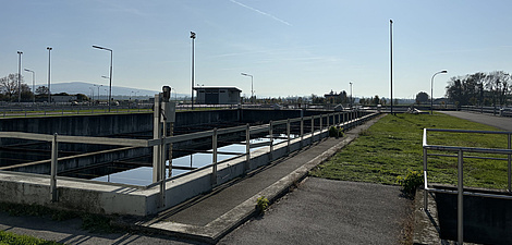 Foto einer Kläranlage. Der Himmel ist blau, darunter ist links ein Becken mit einem Geländer außen herum zu sehen. Rechts ist eine Wiese.