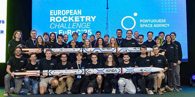 A large group of people stand in two rows, one behind the other. The front and back rows each hold a rocket in front of them.