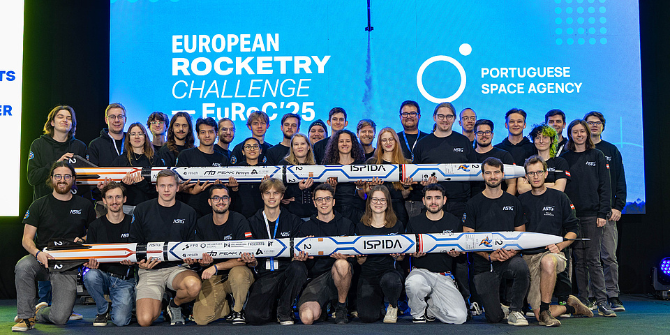 A large group of people stand in two rows, one behind the other. The front and back rows each hold a rocket in front of them.