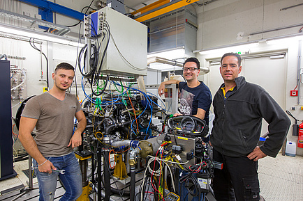 Three men in front of a device with many colourful cables, on the left in a brown T-shirt, in the middle a gentleman with glasses and on the right a gentleman with a grey jacket