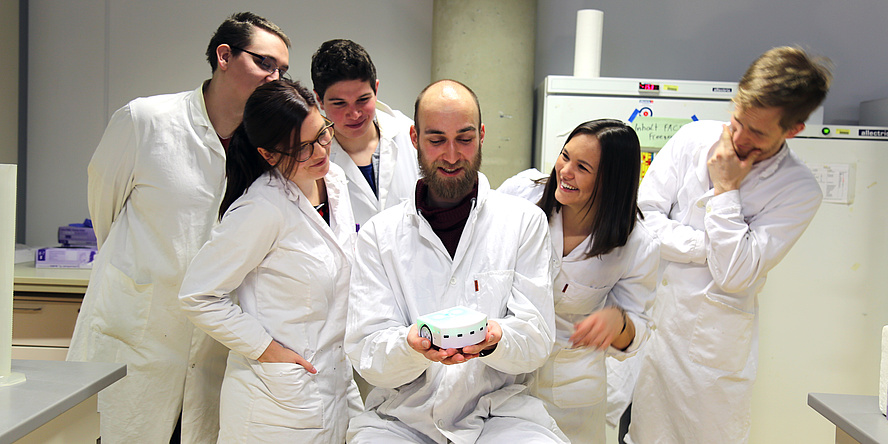 Sechs Mitglieder des aktuellen iGEM-Teams in weißen Kitteln in Laborräumlichkeiten.