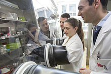 A woman and two men in lab coats. The woman's arms are stuck in black gloves that reach into a glass box.