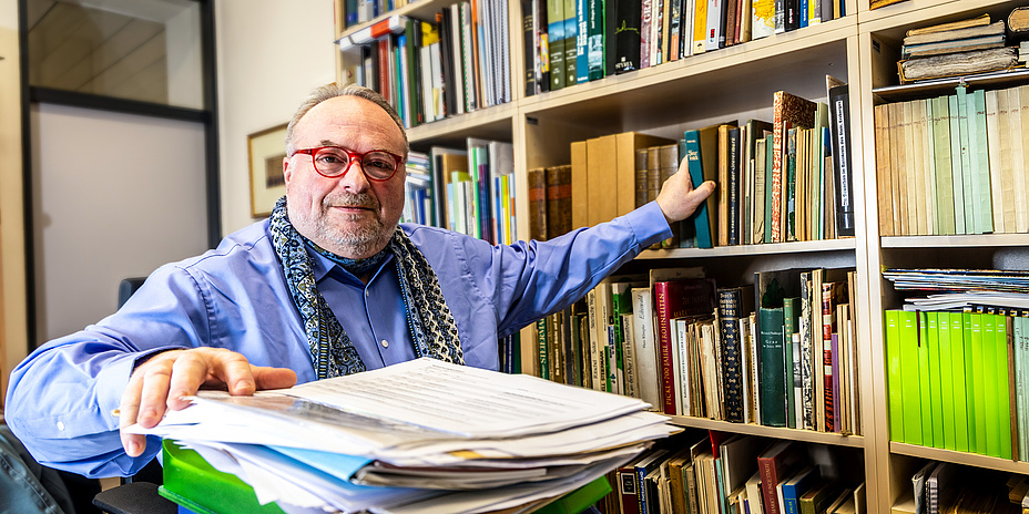 Mann mit roter Brille und Dreitagebart blickt lächelnd in die Kamera. Im Hintergrund sind Bücher und Unterlagen zu sehen.  